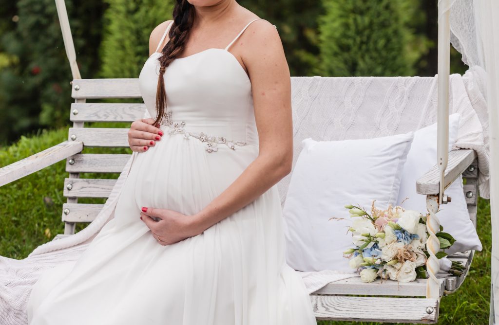 Schwanger heiraten - Braut sitzt auf einer Schaukel und umarmt liebevoll ihren Babybauch