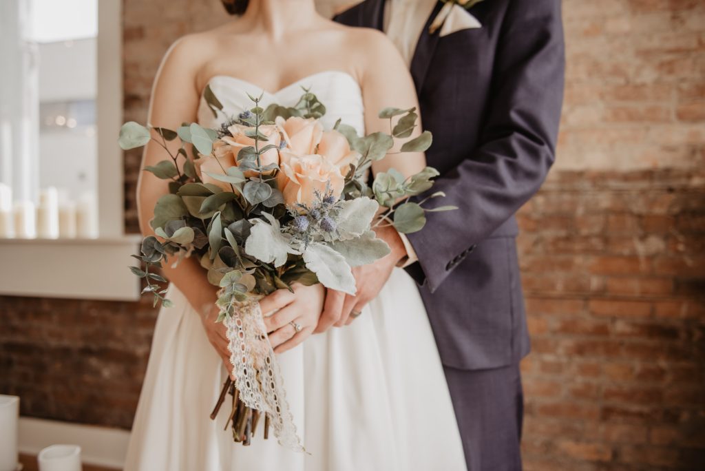 Hochzeitsdienstleister - Braut mit Brautstrauß aus Eukalyptus und Rosen und Bräutigam stehen vor Steinwand in ihrer Traumlocation 