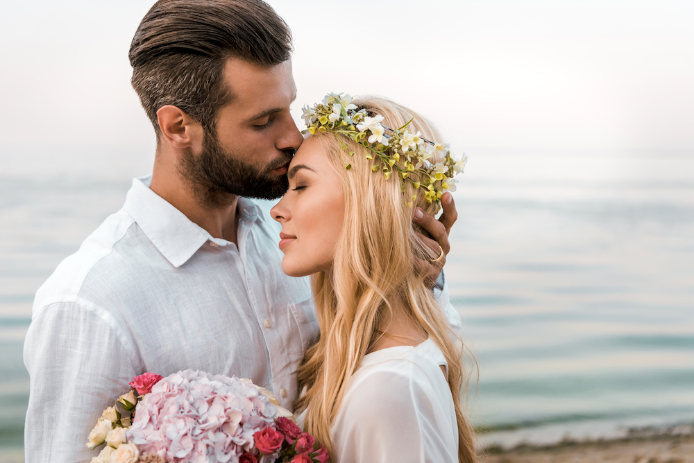 Ehemann küsst Stirn seiner Braut im Sandstrand am Meer