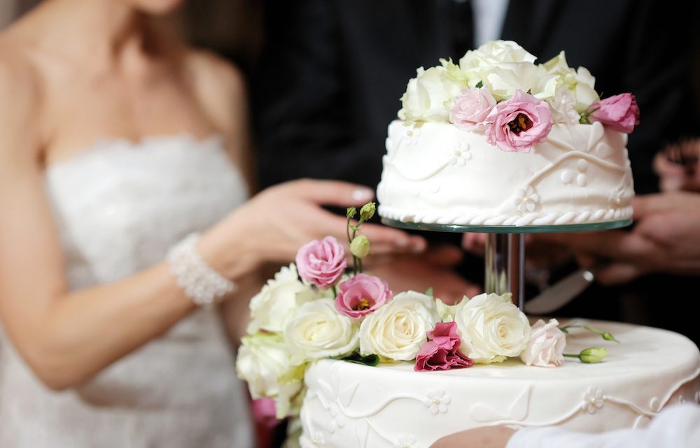 Braut und Bräutigam schneiden gemeinsam die cremefarbene mit Rosen verzierte Hochzeitstorte an