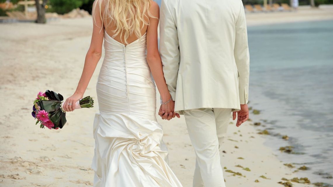 Destination Wedding: Brautpaar geht Hand in Hand am weißen Stand entlang