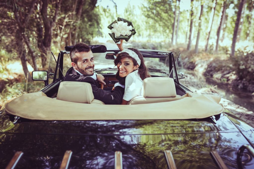 Glückliches Brautpaar fährt mit Cabrio Allee entlang