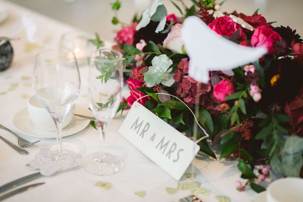 Brauttisch edel dekoriert mit weißem Mr & Mrs Schild und einem Blumenbouquet in den Farben Rot und Grün